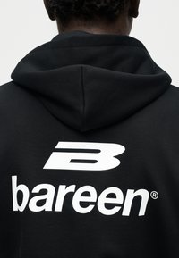 Άτομο που φοράει μαύρο φούτερ με κουκούλα, με λευκό στυλιζαρισμένο λογότυπο "B" και το κείμενο "bareen" στην πλάτη, με την πλάτη γυρισμένη προς την κάμερα.
