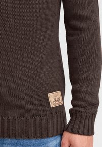 Brun strikket sweater med ribbede manchetter og kant. Har en struktureret overflade og et rektangulært mærke, der viser "Solid" i en vintage stil.