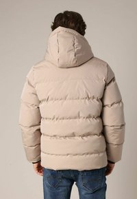 Beige gewatteerd jack met capuchon, uitgevoerd met een doorgestikt ontwerp en een geribbelde zoom. Gecombineerd met blauwe spijkerbroeken, waarbij textuur en constructiedetails zichtbaar zijn.