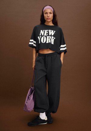 T-shirt nera corta con stampa "NEW YORK", strisce bianche sulle maniche, abbinata a pantaloni della tuta neri larghi e a una borsa a tracolla viola.
