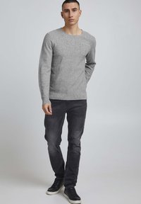 Pull gris texturé à manches longues avec une poche poitrine, associé à un jean foncé et des baskets noires, présentant une coupe ajustée et un col rond.