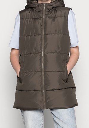 Personne portant une veste sans manches matelassée marron foncé avec fermeture éclair à l'avant et poches, par-dessus une chemise bleu clair à manches courtes et un jean en denim clair.