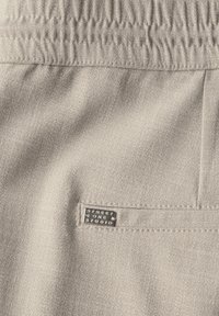 Licht beige stof met een textuurafwerking, voorzien van een elastische tailleband en een rechthoekig label met de tekst "STREET ONE STUDIO."