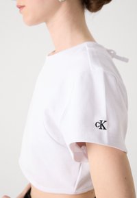 Λευκό κοντό μπλουζάκι από βαμβάκι με κοντό μανίκι. Έχει μαύρο κεντητό λογότυπο 'cK' στον αριστερό μανίκι και λεπτομέρεια δέσιμο στην πλάτη.