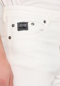 Des jeans en denim blanc sont ornés d'une étiquette noire portant la mention "VERSACE JEANS COUTURE." Détail avec du matériel en métal argenté et des coutures claires.