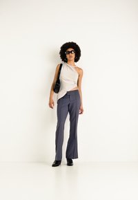 Top grigio chiaro a spalla sola, pantaloni a campana blu navy, borsa nera e occhiali da sole, con una texture di tessuto liscia. Modella posiziona su uno sfondo bianco.