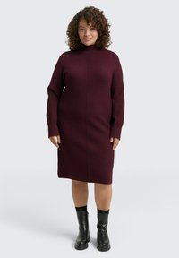 Bordeauxfarbendes Strickkleid mit hohem Kragen und langen Ärmeln; hat eine taillierte Form und gerippte Bündchen. Getragen mit schwarzen Stiefeletten.