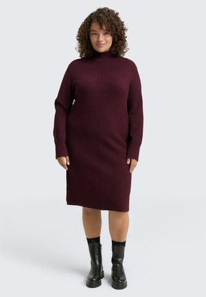 Robe tricotée bordeaux avec un col montant et des manches longues ; présente une coupe ajustée et des poignets côtelés. Portée avec des bottines noires.
