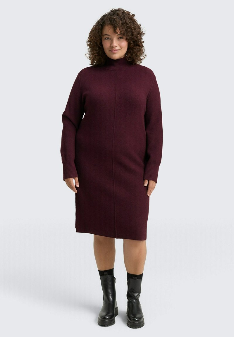 Bordeauxfarbendes Strickkleid mit hohem Kragen und langen Ärmeln; hat eine taillierte Form und gerippte Bündchen. Getragen mit schwarzen Stiefeletten.