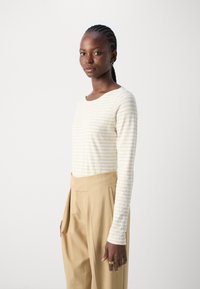 Un top striped bej și alb cu mâneci lungi, asortat cu pantaloni bej cu talie înaltă. Topul are un decolteu rotund și mâneci ajustate.