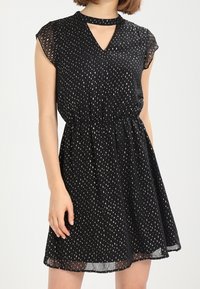 Femme portant une robe noire à manches courtes avec des motifs en forme de goutte d'argent et un décolleté en trou de serrure, se tenant devant un fond blanc.