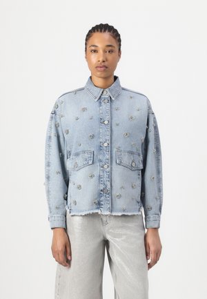 Femeie cu părul împletit poartă o jachetă de denim albastru deschis decorată cu mici ornamente în formă de trandafir și pantaloni argintii, stând pe un fundal alb.