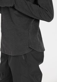 Schwarzes Langarmshirt mit abgerundetem Saum und weicher Textur, kombiniert mit schwarzen Hosen, die eine Reißverschlusstasche und eine glatte Oberfläche aufweisen.