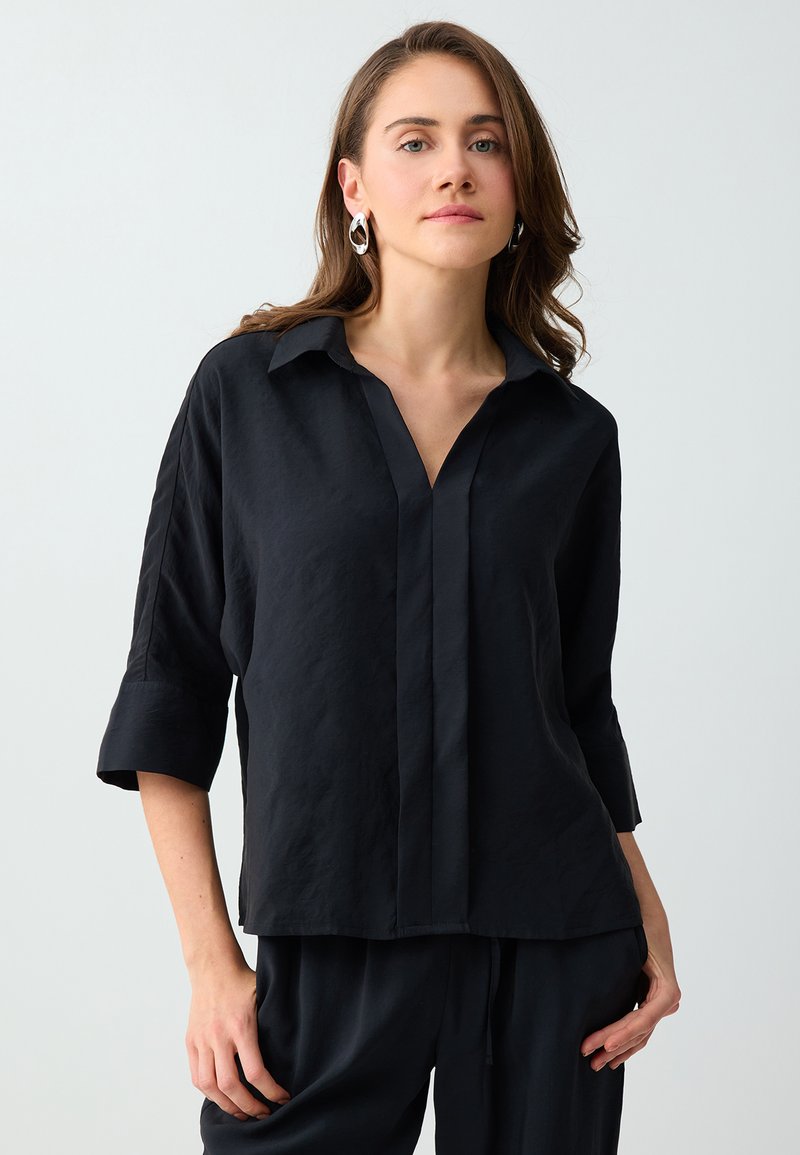 Jeune femme aux cheveux ondulés, lâchés, portant des boucles d’oreilles argentées en anneaux et une chemise noire à manches trois-quarts, debout devant un fond uni.