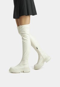 Botas blancas hasta el muslo, confeccionadas en material suave, con una suela gruesa y cremallera lateral, combinadas con una falda negra de rayas.