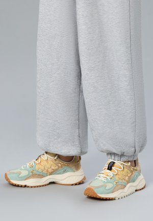 Persona con pantalones deportivos grises y zapatillas multicolores beige, verdes y marrones con cordones blancos, de pie sobre un suelo gris.