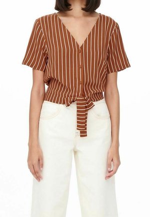 Vrouw die een bruin, kort mouwtjes, V-hals, gestreept overhemd met witte verticale strepen draagt, dichtgeknoopt in de taille, gecombineerd met witte, hooggetailleerde broek.