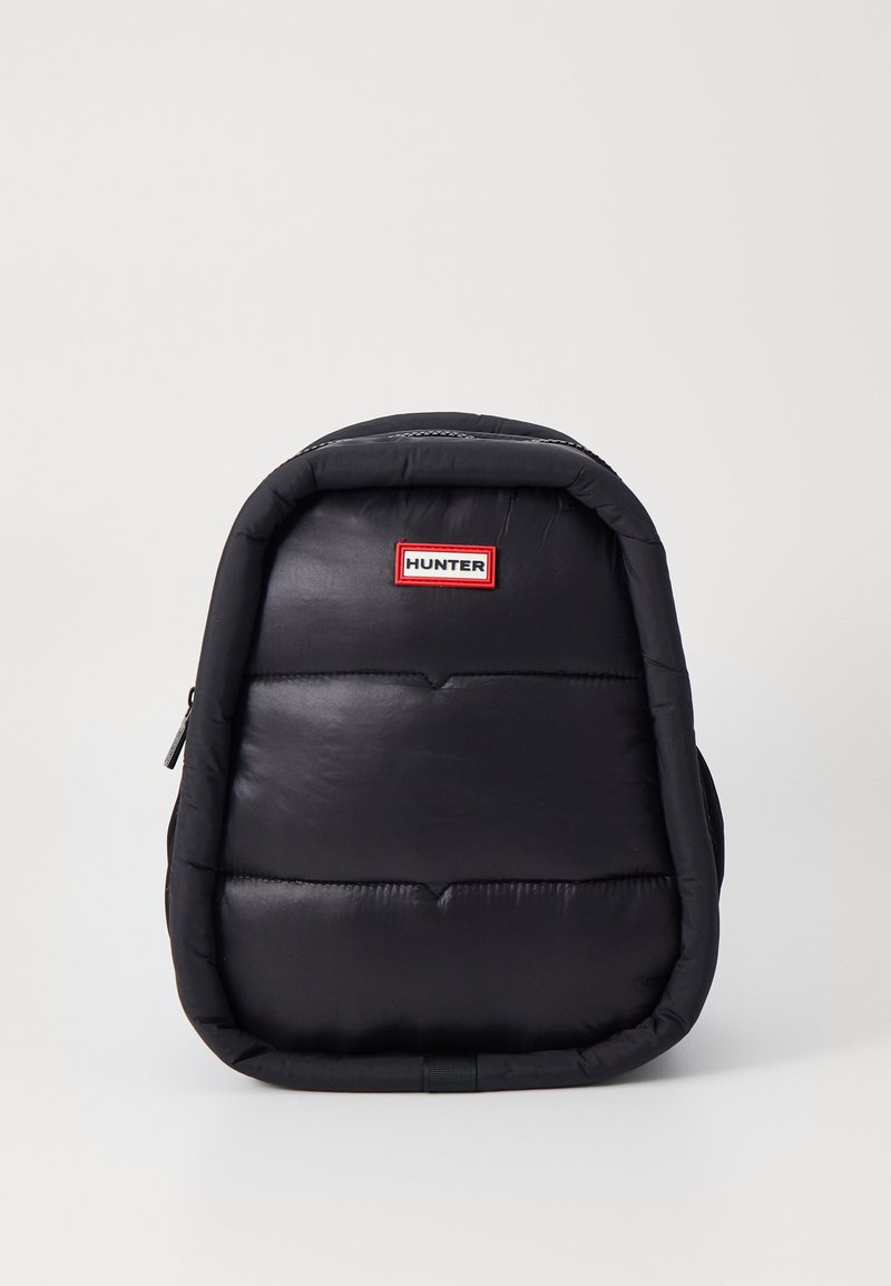 Mochila negra acolchada con forma rectangular, cremallera frontal y un parche de logo con ribete rojo. Texture suave con un diseño minimalista.