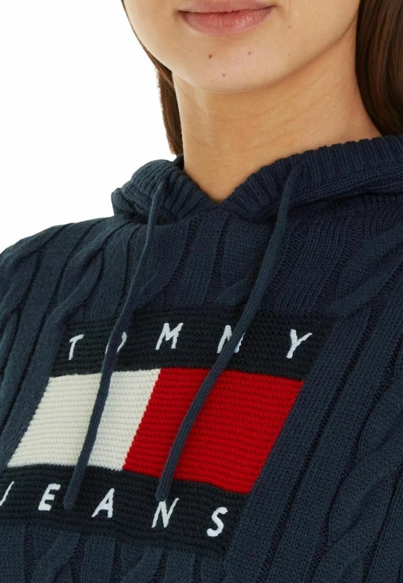 Personne portant un sweat à capuche tricoté bleu marine avec un grand logo rectangulaire rouge, blanc et noir Tommy Jeans sur la poitrine.