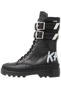 Černá kožená šněrovací bota se dvěma stříbrnými přezkami, strukturovanou gumovou podrážkou a bílým grafickým detailem "KARL" na boku. Masivní design.