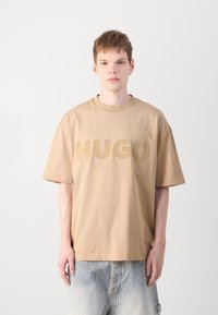 Tricou din bumbac bej, cu o croială oversized, cu mâneci scurte și logo-ul embosat "HUGO" pe față. Purtați cu pantaloni scurți din denim deschis.