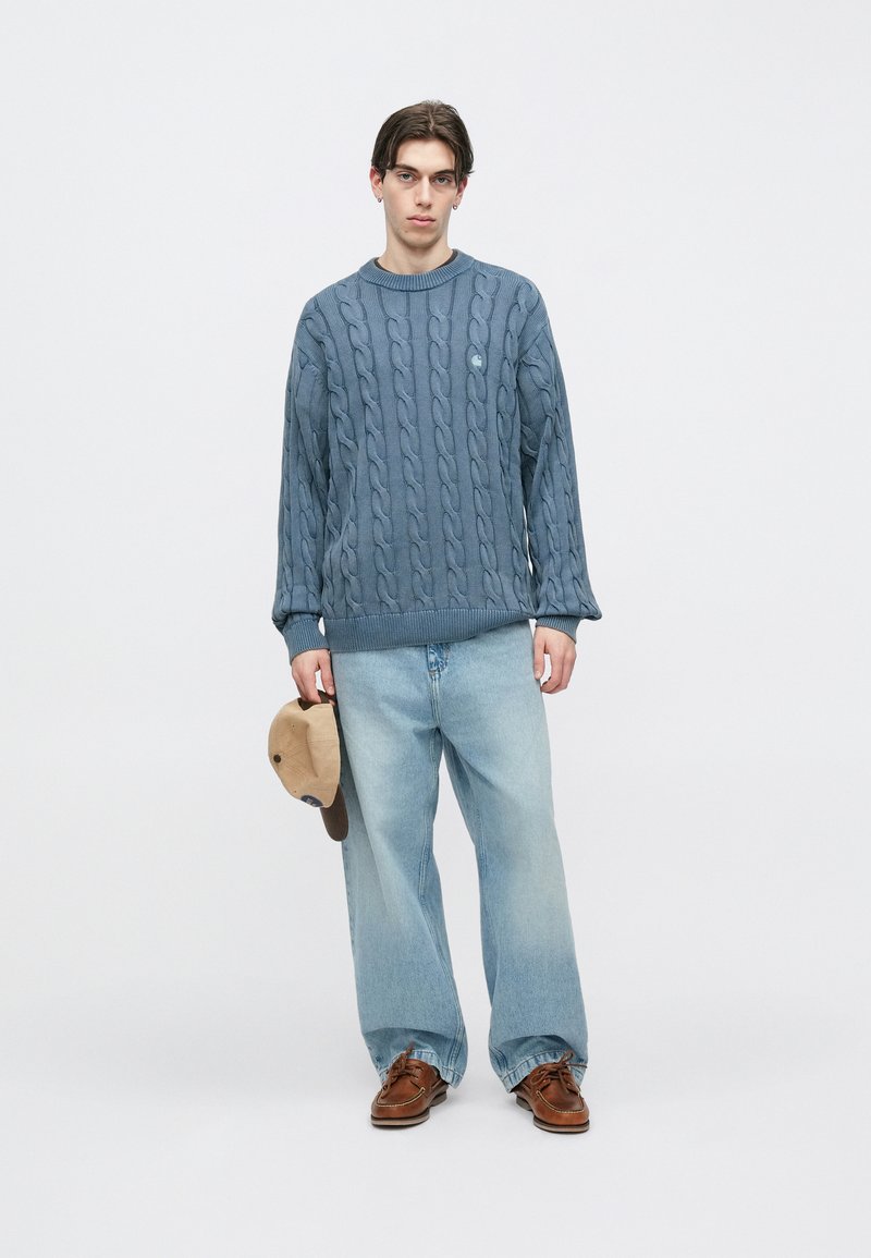 Blauwe kabelgebreide trui met lange mouwen, gecombineerd met lichtblauwe wijde jeans. Houdt een beige pet vast, bruine schoenen maken de look compleet.