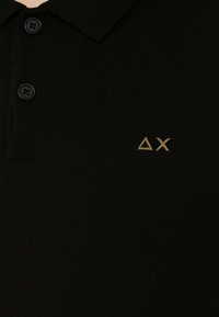 Polo shirt nera in maglia con colletto, due bottoni neri e un logo AX diagonale dorato sulla parte sinistra del petto.