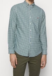 Zelená a bílá kostkovaná košile na knoflíky s dlouhými rukávy a límečkem; na hrudi je malé logo. Nositelná s khaki kalhotami.