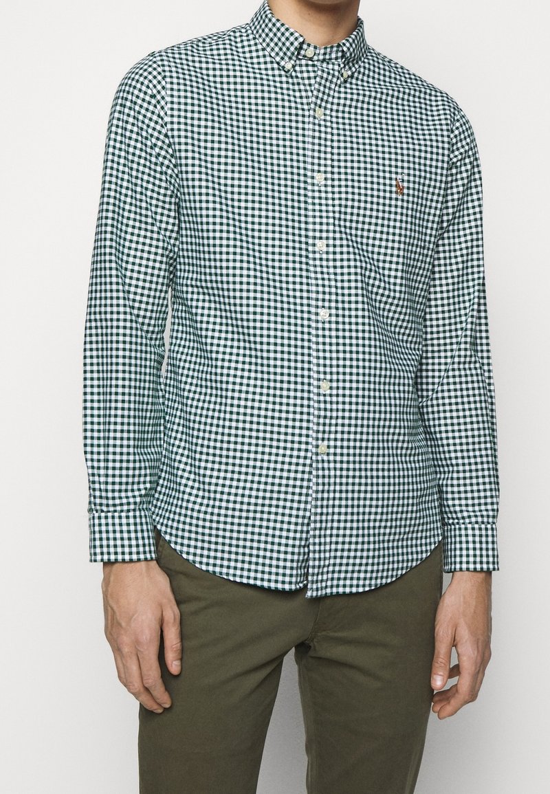 Zelená a bílá kostkovaná košile na knoflíky s dlouhými rukávy a límečkem; na hrudi je malé logo. Nositelná s khaki kalhotami.