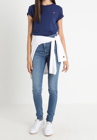 Dunkelblaue Baumwoll-T-Shirts mit umgeschlagenen Ärmeln, hellblaue Skinny-Jeans, ein weißer Pullover, der um die Taille gebunden ist, und weiße Sneakers.