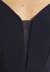 Gros plan d'une robe bleu marine à décolleté en V profond avec une partie en mesh transparent et une peau visible sur le haut de la poitrine présentant un petit grain de beauté.