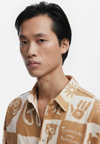 Kortærmet, knap-op skjorte med et tan- og cremefarvet abstrakt mønster, der fremhæver grafiske designs og tekstureret stof. Med krave og løs pasform.