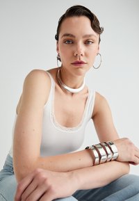 Balta topukas su nėriniu, sidabrinė chokero apyrankė ir kelios sidabrinės apyrankės. Modelis su trumpais plaukais, šukuotais atgal, sėdi prieš šviesų foną.