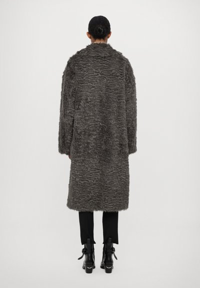 Ilgas, pilkas kailinis paltas su tekstūruotu raštu, turintis apykaklę ir laisvą siluetą. Dėvimas ant juodų kelnių ir aukštakulnių aulinukių.