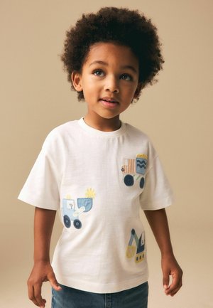 Kind mit lockigem Haar, trägt ein weißes T-Shirt mit bunten Aufnähern von Baumaschinen auf beigem Hintergrund.