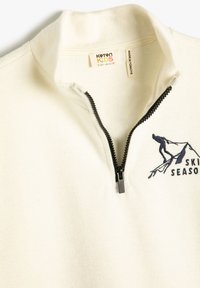 Kremowy sweter dziecięcy z czarnym zamkiem błyskawicznym do jednej czwartej, z haftowanym narciarzem i napisem "SKI SEASON" na klatce piersiowej, widoczna metka Koton Kids.