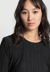 Top preto com nervuras e decote redondo, apresentando um padrão texturizado. O tecido parece suave, com um brilho sutil e um design de ajuste slim.