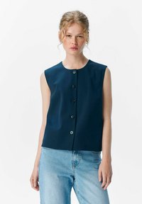 Débardeur bleu marine sans manches avec un col rond, fermeture à boutons sur le devant et une coupe légèrement décontractée. Associé à un jean en denim bleu clair.
