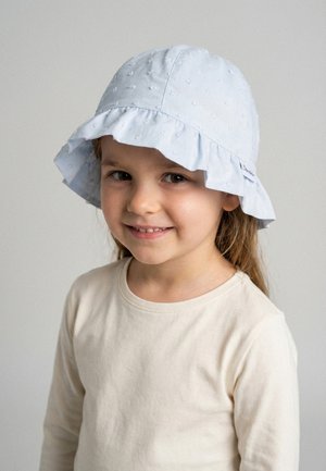 Lächendes junges Mädchen mit langem Haar, das einen hellblauen, gerüschten Sonnenhut und ein cremfarbenes Langarmshirt vor einfarbigem Hintergrund trägt.