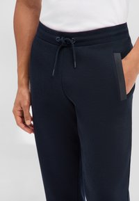 Mężczyzna ubrany w granatowe spodnie dresowe z paskiem ściągającym w talii i kieszenią ozdobioną czarną lamówką, z ręką spoczywającą blisko kieszeni, w białej włożonej koszuli.