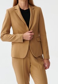 Blazer in kameelkleur met een getailleerd ontwerp, met ingestikte revers en een enkele borstzak. Gemaakt van een gladde stof, gecombineerd met een zwart topje.