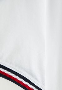 Biała koszulka polo z tkaniny o fakturowanym wzorze, z dołem ozdobionym czerwonymi i granatowymi paskami, które podkreślają detale projektu.