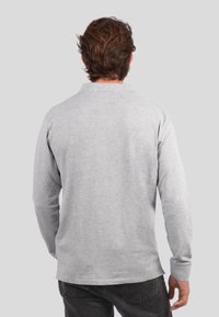 Mies, jolla on ruskeat hiukset, pukeutuneena vaaleanharmaaseen pitkään hihapaitaan ja tummiin farkkuihin, selin yksinkertaista harmaata taustaa vasten.