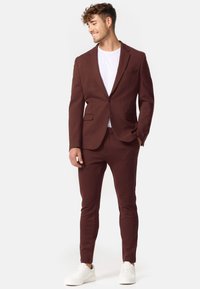 Bordeauxfarbener Anzug mit tailliertem Jacket und schmal geschnittenen Hosen, kombiniert mit einem weißen T-Shirt und weißen Sneakers. Glatte Stofftextur.