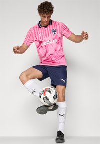 Rožinė futbolo marškinė su purslų raštu ir juodais akcentais, derinama su tamsiai mėlynomis šortais ir baltomis kojinėmis. Žaidėjas balansuoja ant futbolo kamuolio.