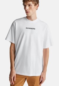 Mladý muž v bílém tričku s nápisem "ČÍSLA", hnědých kalhotách a tetováním motýla na levém předloktí, dívá se do strany.