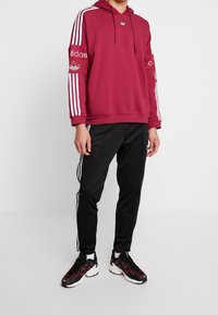 Kastanjebrunt hoodie med vita ränder på ärmarna, med en liten logotyp. Svarta byxor med vita sidostripes. Svarta sneakers med röda detaljer.