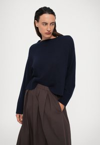 Vrouw in een marineblauwe trui en losse bruine broek met één hand in de zak, staande tegen een effen lichte achtergrond.