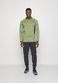Groene langemouwen top met textuurstof, donkere zijkanten en ronde halslijn. Gecombineerd met zwarte tapered broek en zwart/blauwe sportschoenen.