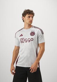 Hvid fodboldtrøje med maroon detaljer, der har et geometrisk mønster, korte ærmer, rund halsudskæring og sponsorlogoet "Ziggo."
